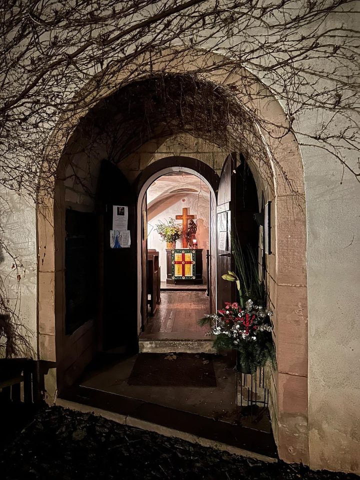 Nachts durch einen berankten Torbogen in die beleuchtete Kapelle mit Kreuz und Altar