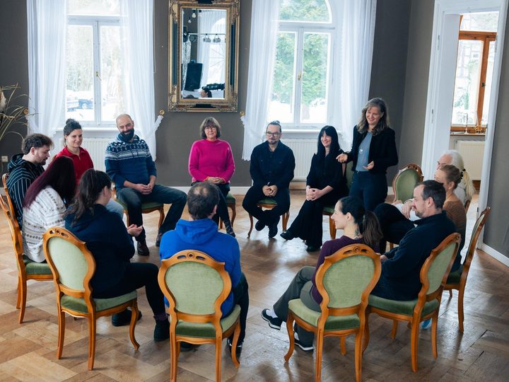 Seminarrunde im hellen Salon: vierzehn Menschen im Stuhlkreis, Goldspiegel und hohe Fenster