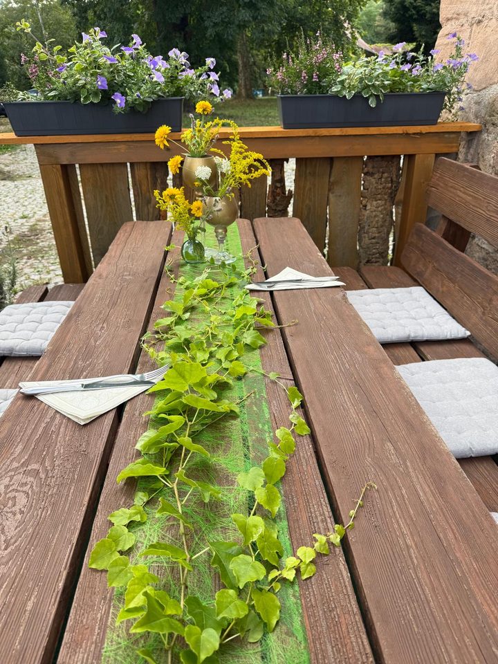 Gedeckte Tafel im Biergarten mit Efeuläufer, Wiesenblumen in Vasen und Sitzkissen auf den Bänken