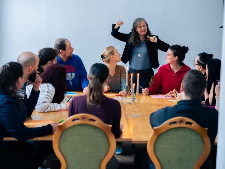 Workshop an der langen Tafel, eine Moderatorin steht und erklärt, Kerzen und Stifte auf dem Tisch