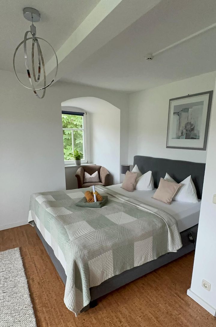 Doppelbett mit grün-weißer Tagesdecke, daneben ein Sessel in der Fensternische